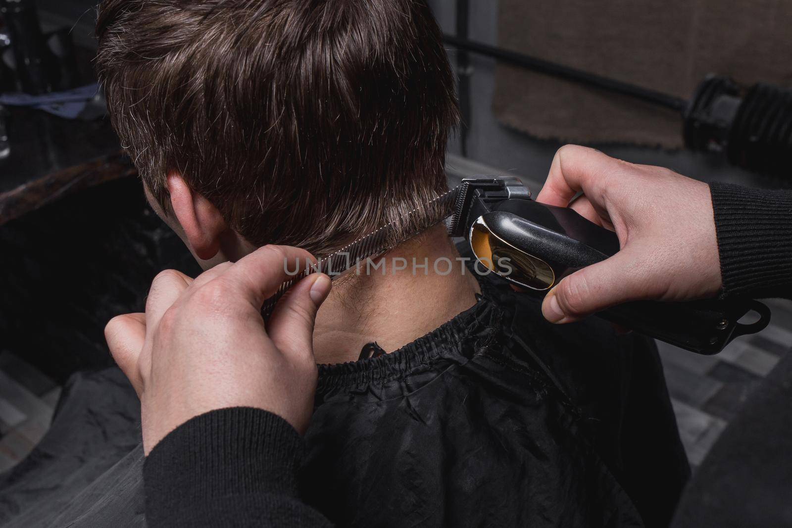 The hands of a barber or hairdresser cut the back of the client's head with dark hair with an automatic machine and comb in the barbershop. Hairdressing.