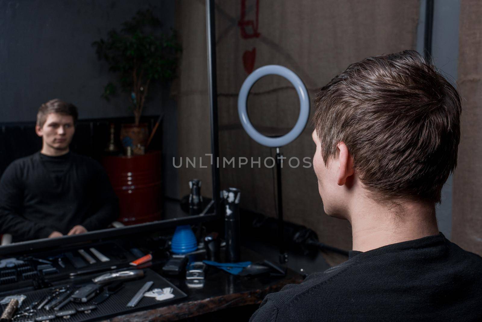 A shaggy guy with long dark hair sits in a chair in a barbershop opposite the reflection in the mirror. The need for hairdressing services.