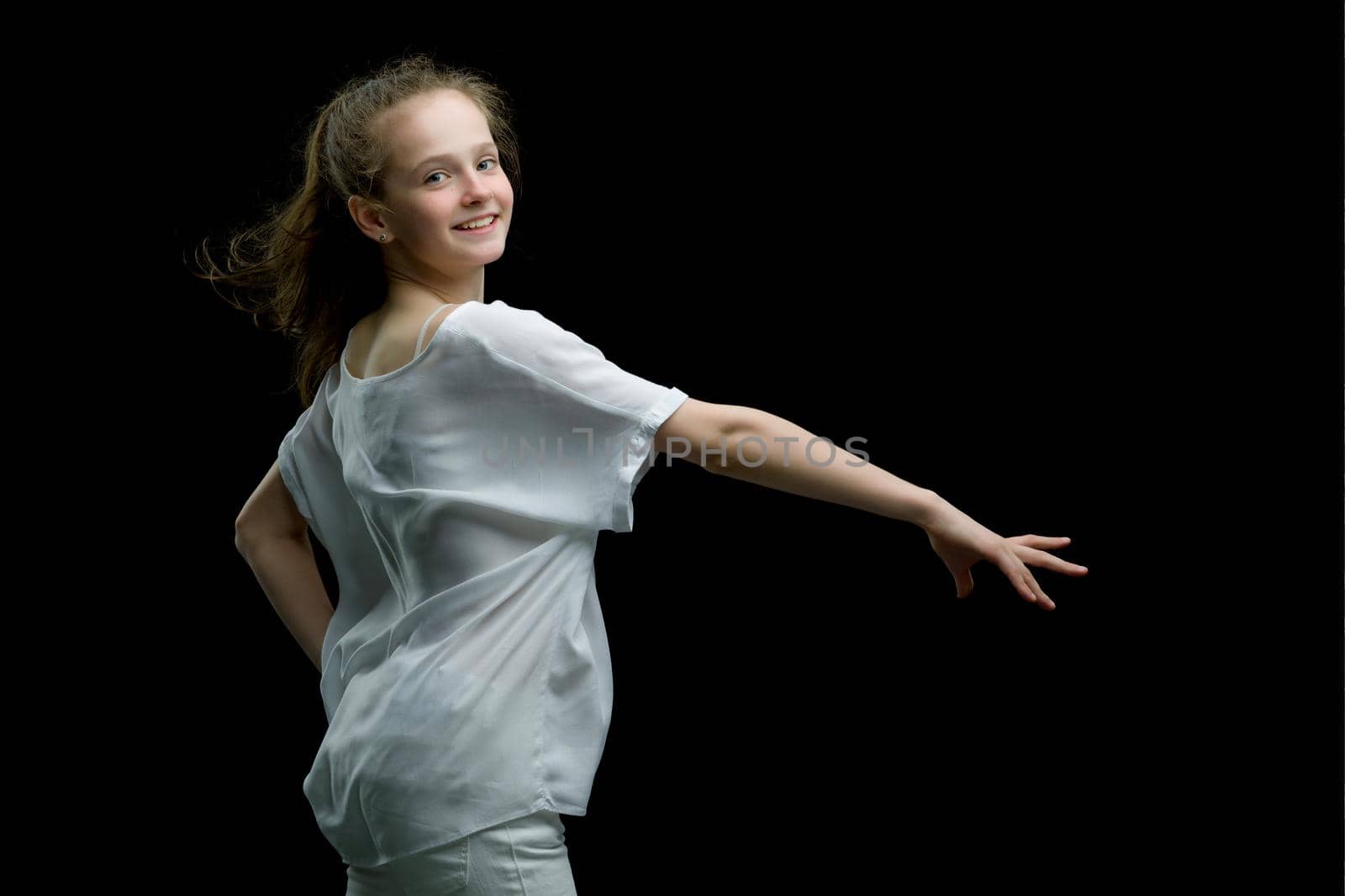 A strong wind blows on the face and clothes of a beautiful little girl of school age. Hair and clothes are fluttering in the wind, and the girl is happy about it. On a black background. Studio photo session