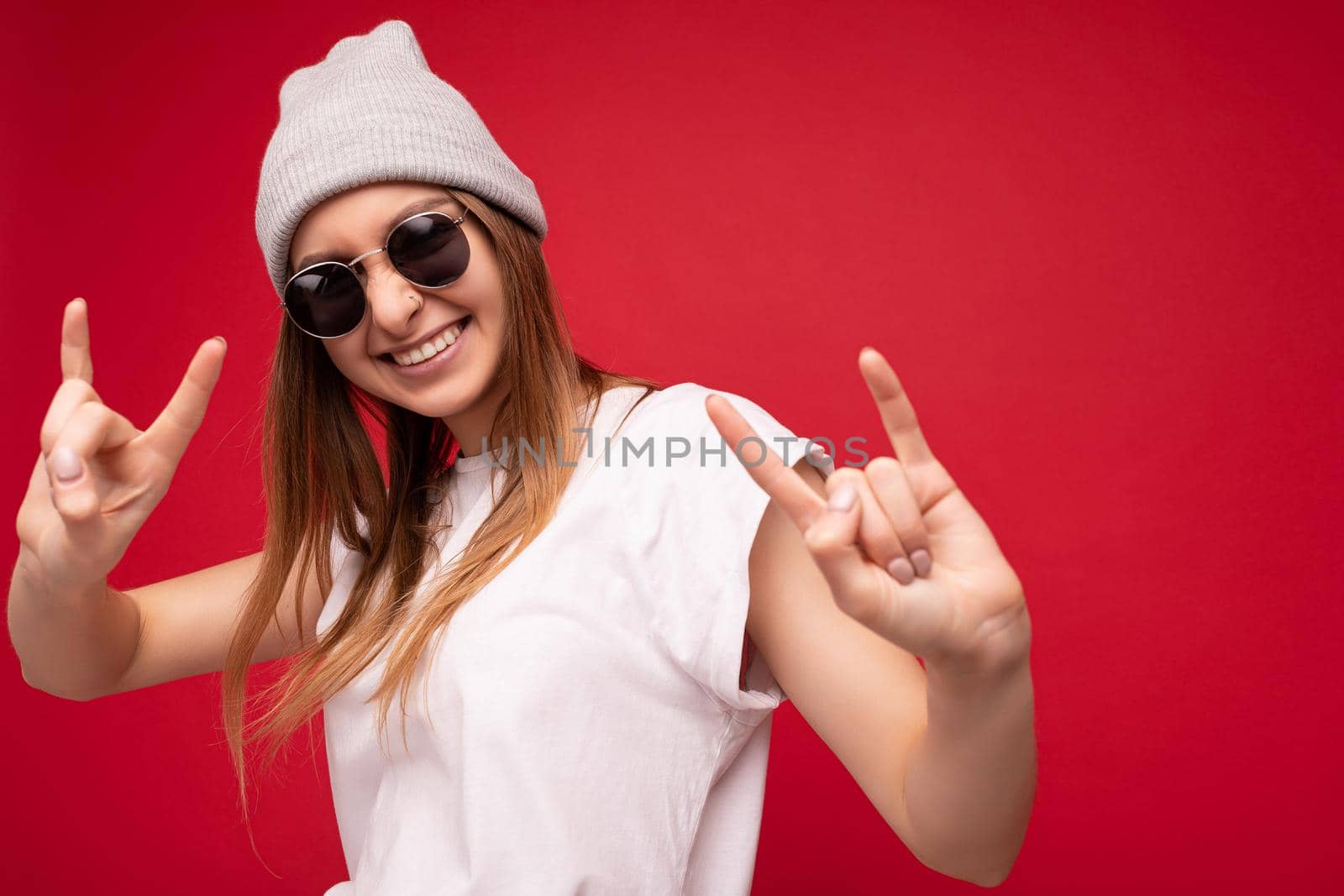 Closeup photo shot of beautiful happy young dark blonde woman isolated over red background wearing casual white t-shirt for mockup and stylish sunglasses looking at camera and showing rock and roll sign.