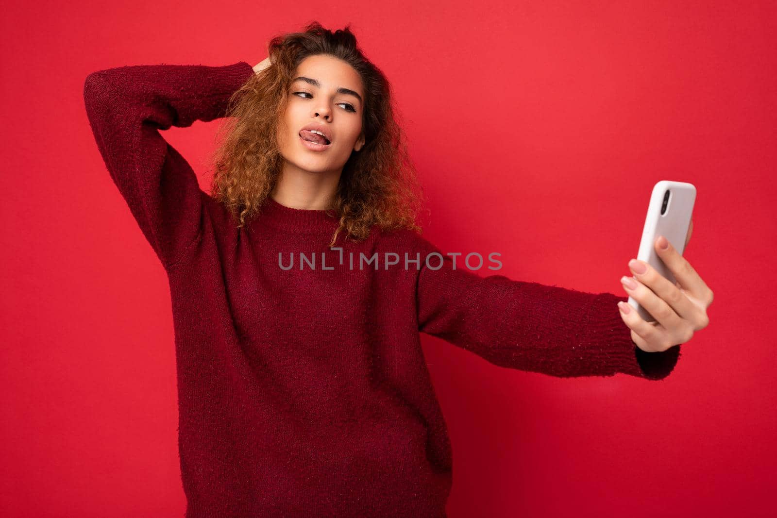 Sexy Beautiful young woman with curly hair wearing dark red sweater isolated on red background wall holding and using smart phone looking at telephone screen and taking selfie and showing tongue.
