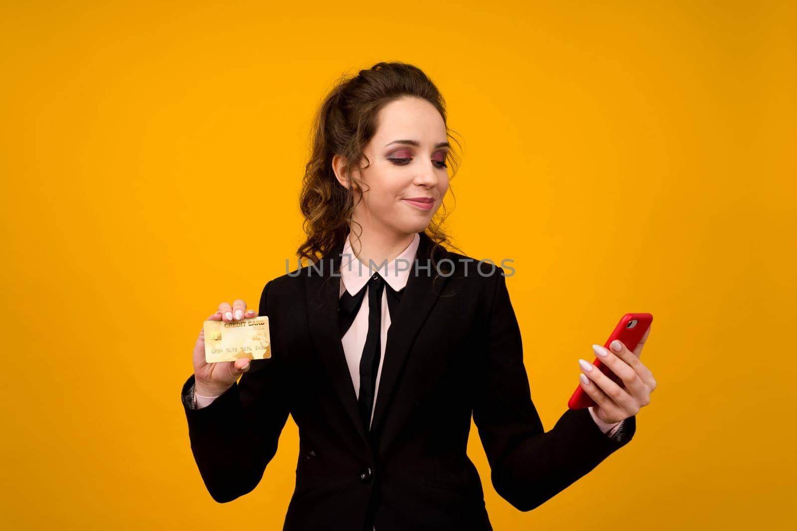 Photo of pleased young woman posing isolated over yellow wall background using mobile phone holding debit card. - image