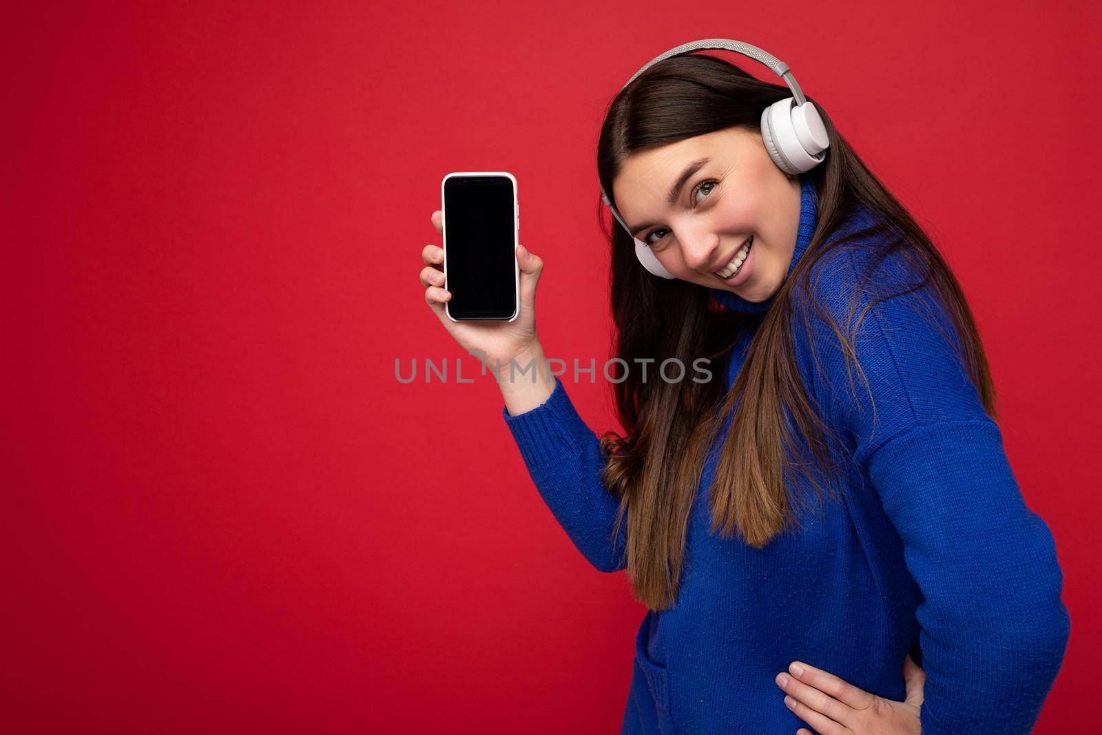 Side-profile portrait photo shot of charming happy young brunette woman wearing casual blue sweater and white earphones isolated over red background with empty space holding in hand mobile phone and showing smartphone with empty screen for mockup listening to music looking at camera.