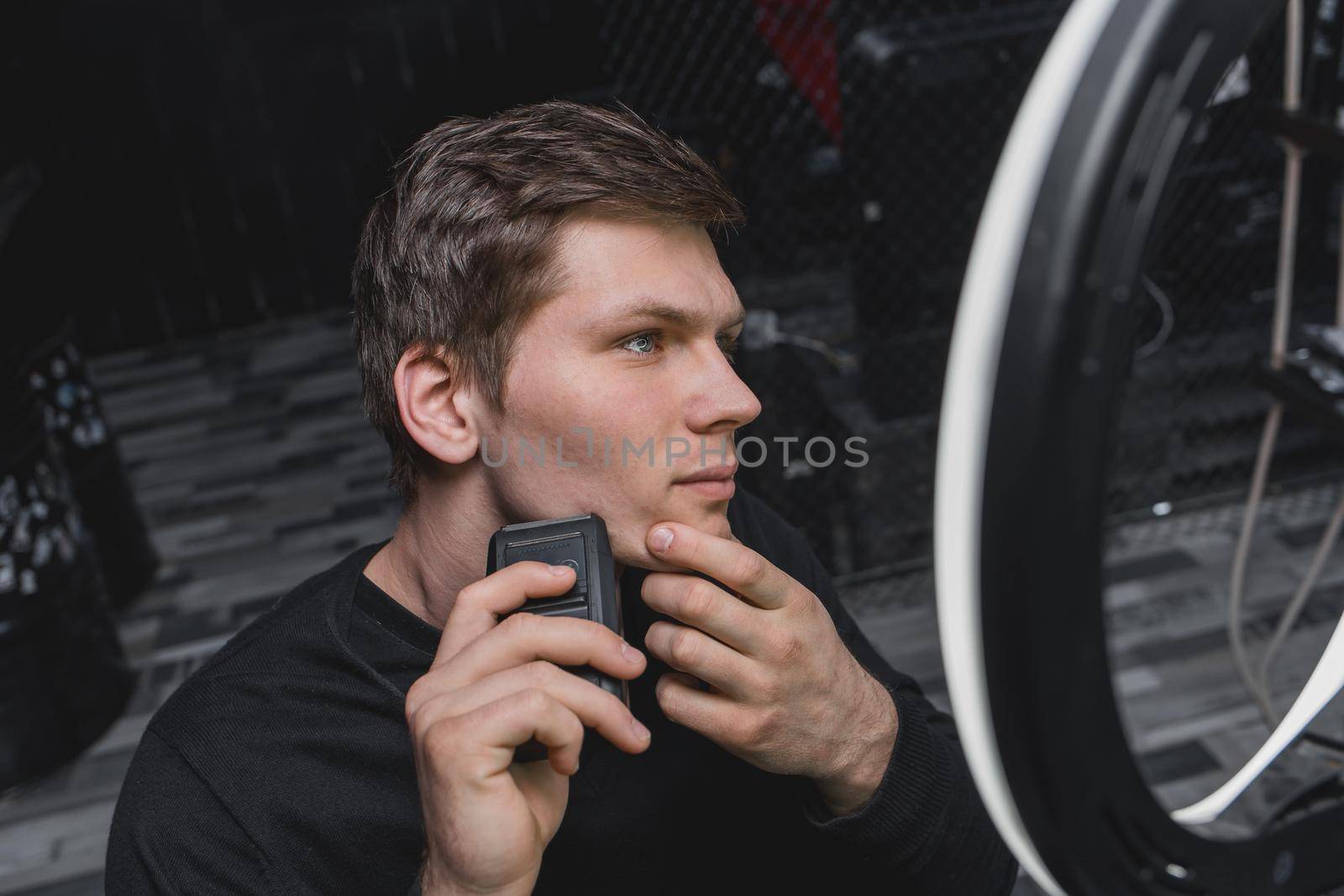 A serious young guy of Caucasian appearance with dark hair shaves with an automatic razor next to the ring lighting. Care for your appearance.