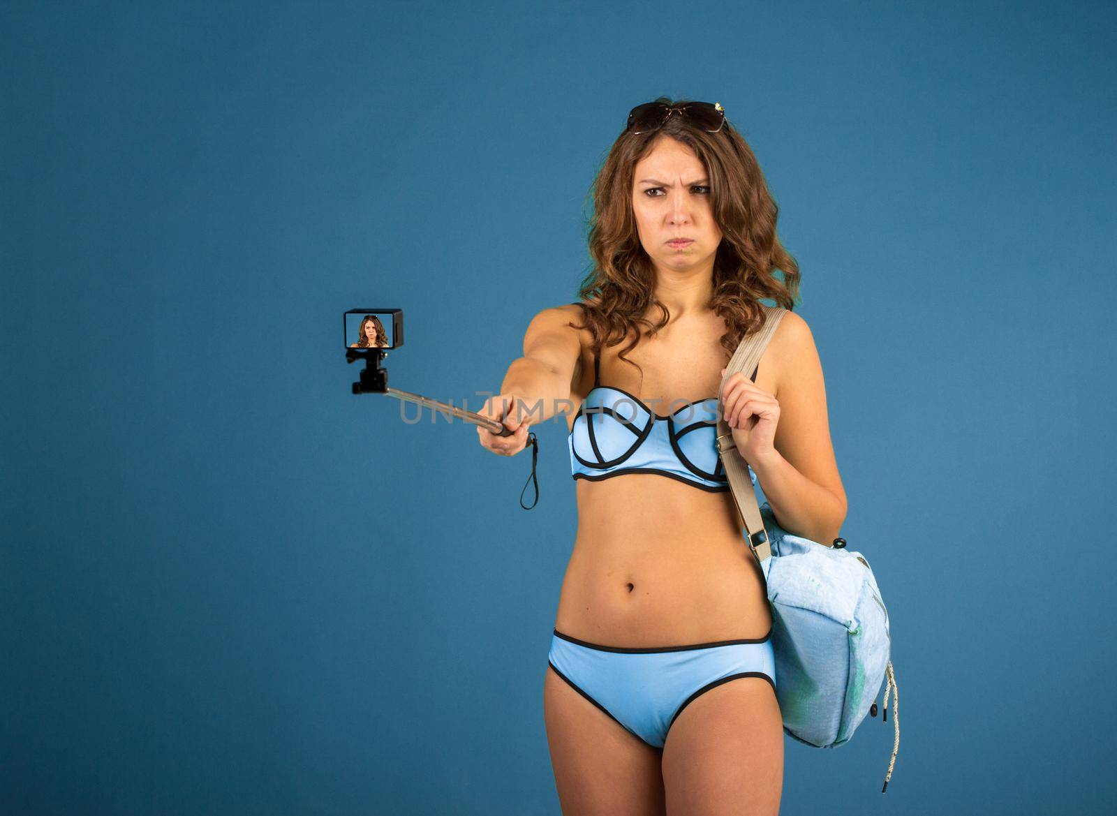 Hipster girl making selfie with action camera on blue background wearing swimwear.