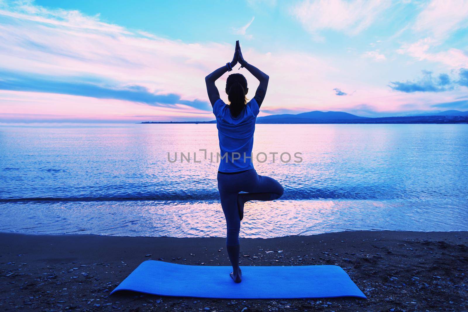 Woman doing yoga exercise in pose of tree on beach near the sea in summer in the morning, rear view