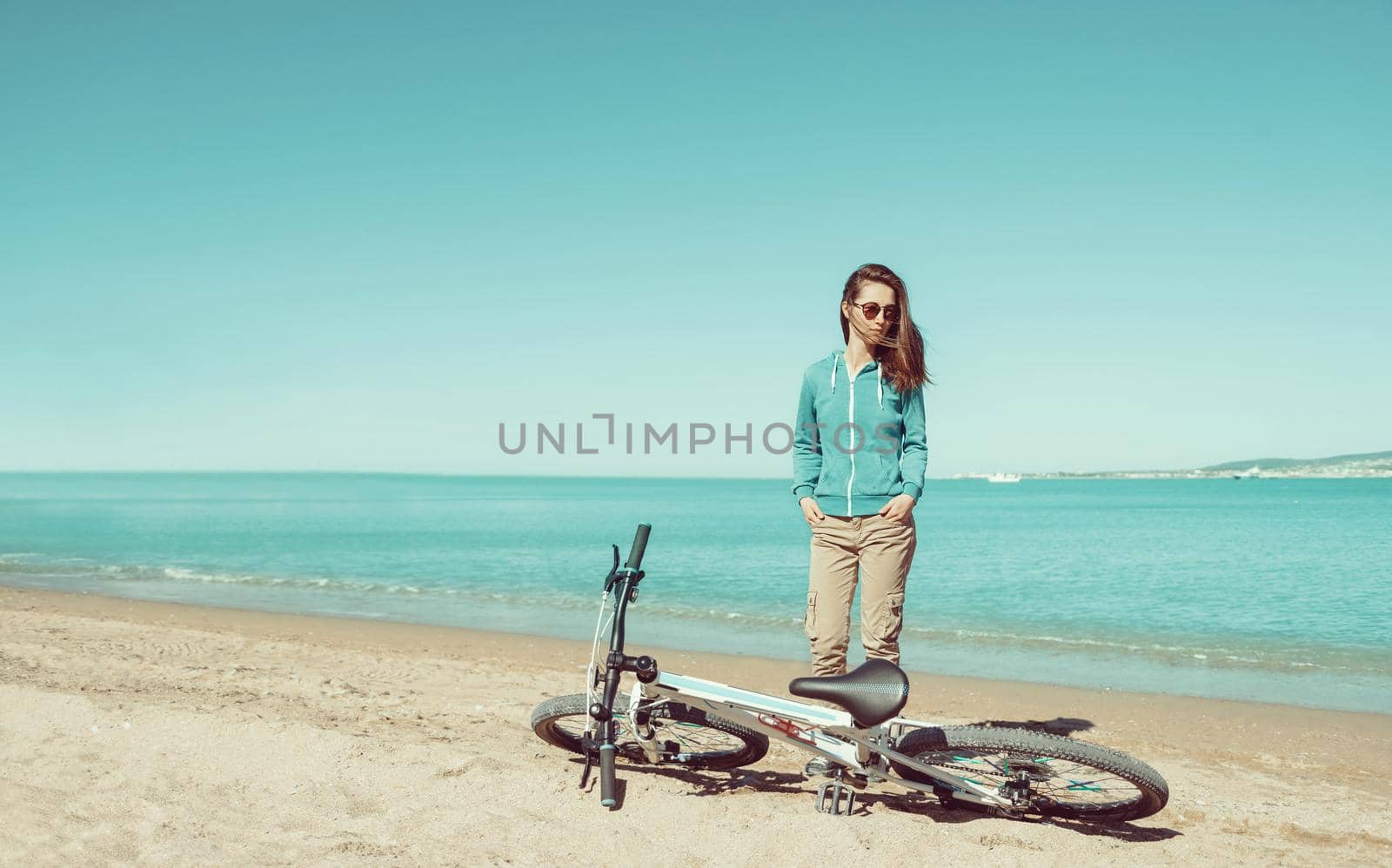 Beautiful young woman in sunglasses standing near a bicycle on sand beach in summer