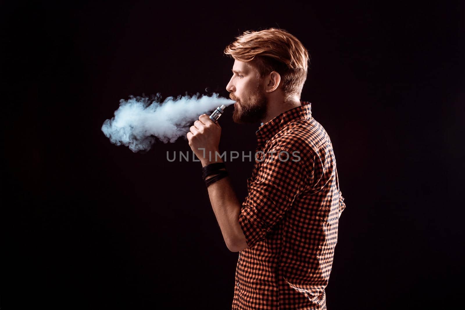 young man wearing a plaid shirt smokes an electronic cigarette on a black background