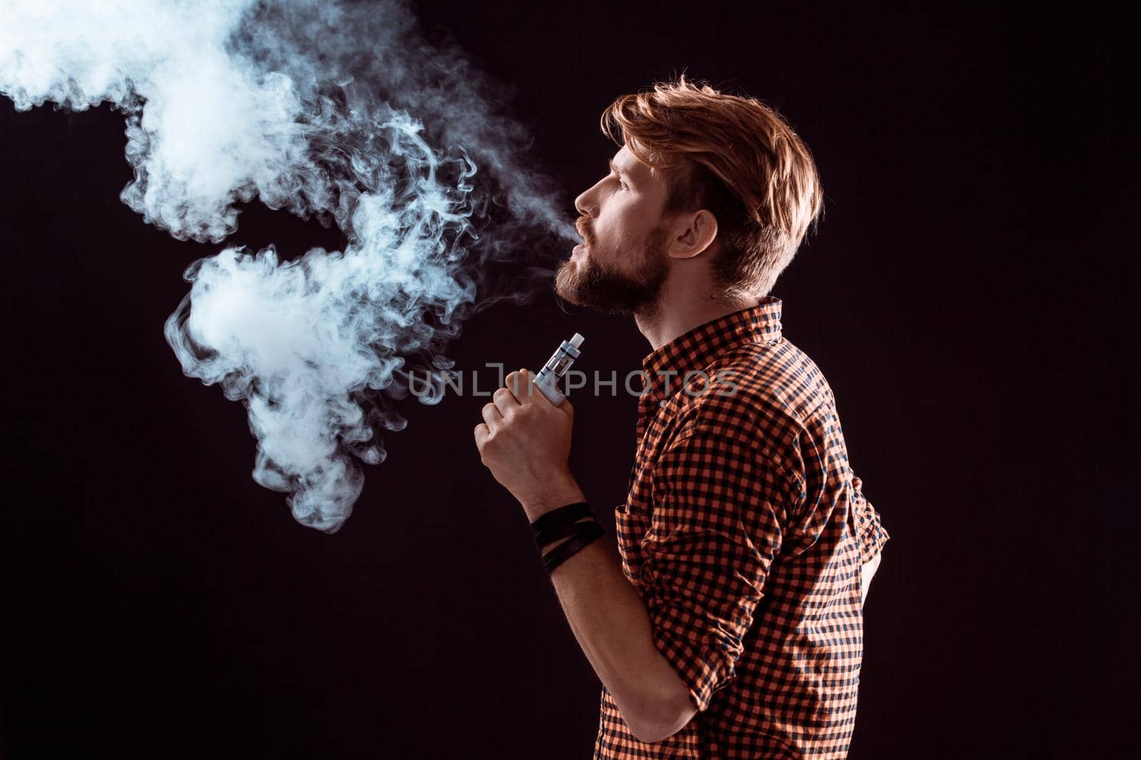 young man wearing a plaid shirt smokes an electronic cigarette on a black background