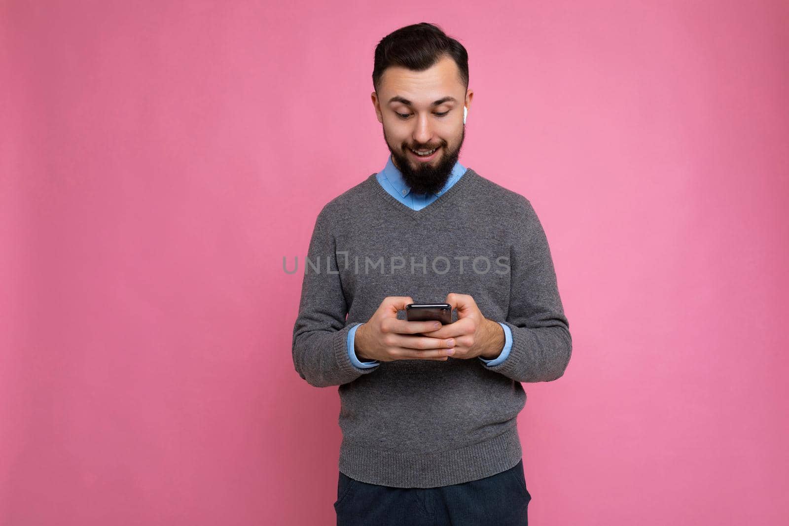 Photo shot of surprised cool good looking brunet unshaven young man with beard wearing grey sweater and blue shirt isolated over pink background wall with empty space holding in hand and using mobile phone messaging sms online looking down at device display.