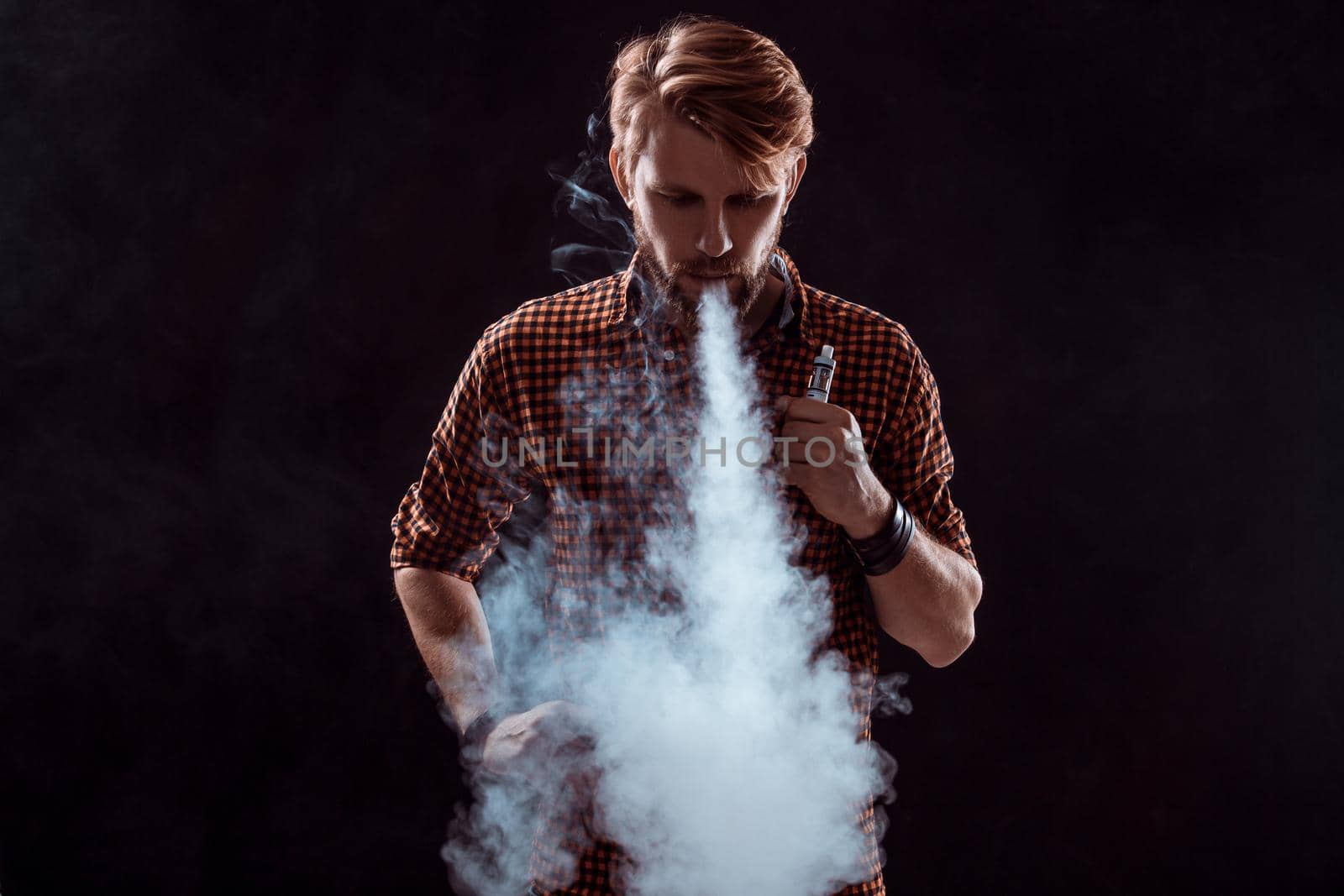 young man wearing a plaid shirt smokes an electronic cigarette on a black background