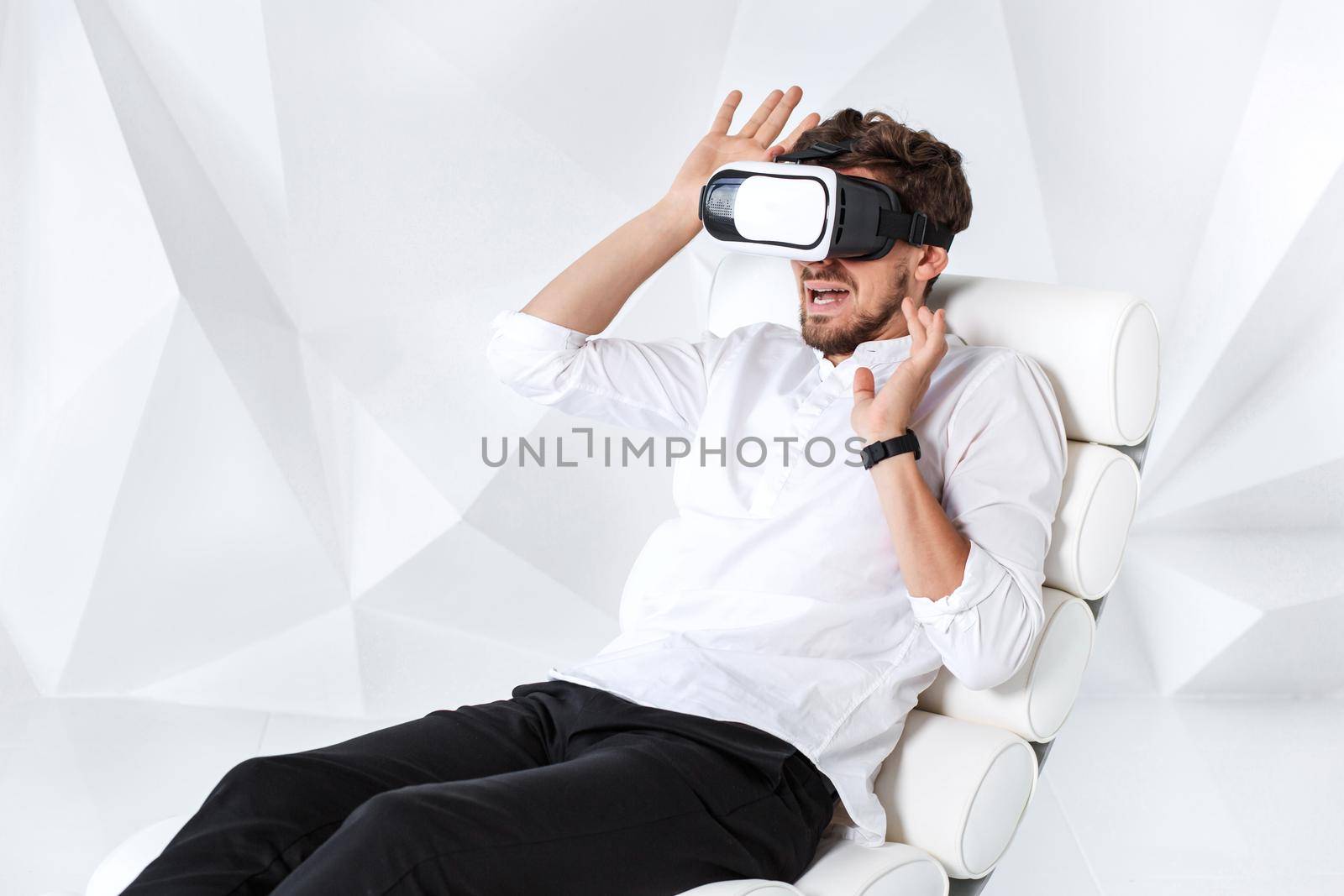 Excited young man is getting experience using VR-headset glasses of virtual reality gesticulating with his hands. A young man sits on a comfortable armchair in a room with white walls