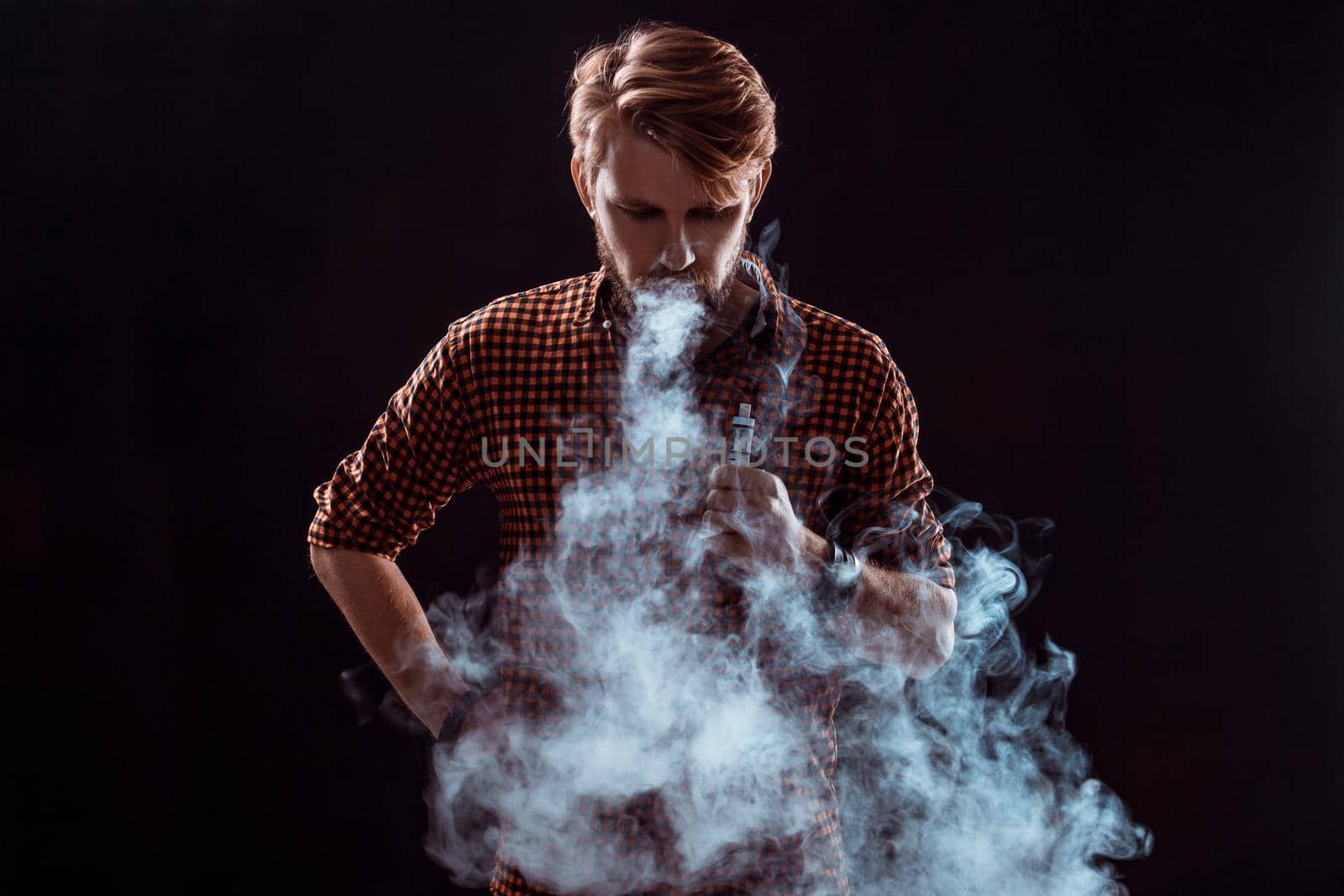 young man wearing a plaid shirt smokes an electronic cigarette on a black background