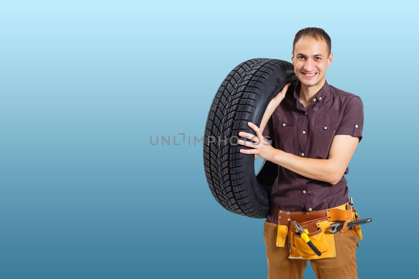 car mechanic carrying tire on blue background. happy man smiling amd looking into camera