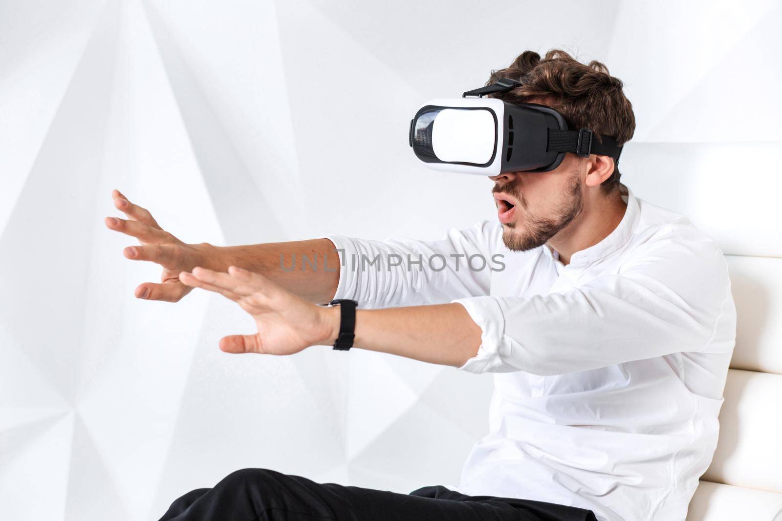 Excited young man is getting experience using VR-headset glasses of virtual reality gesticulating with his hands. A young man sits on a comfortable armchair in a room with white walls