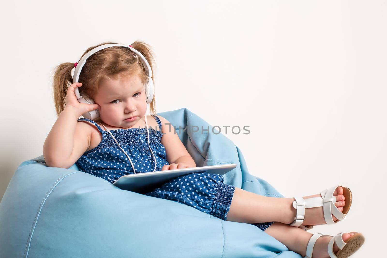 Cute little girl in headphones listening to music using a tablet and smiling while sitting on blue big bag. On white background.