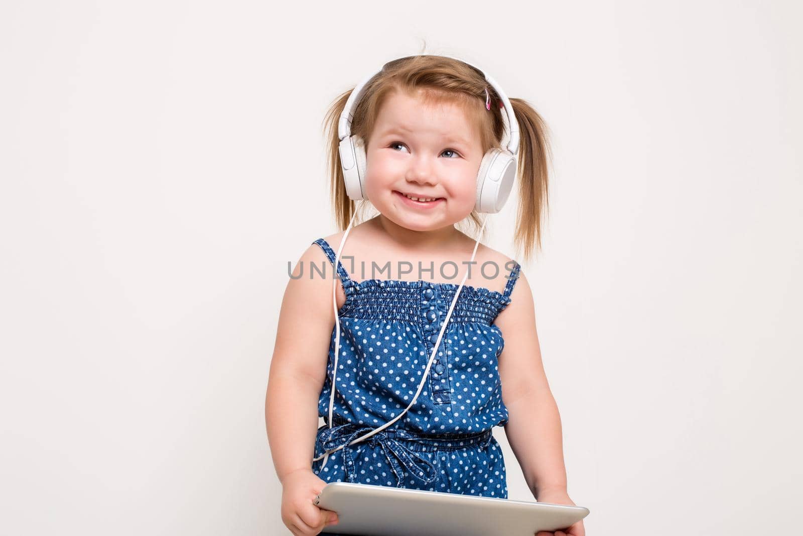 Cute little girl in headphones listening to music using a tablet and smiling on white background. The child does not look at the camera