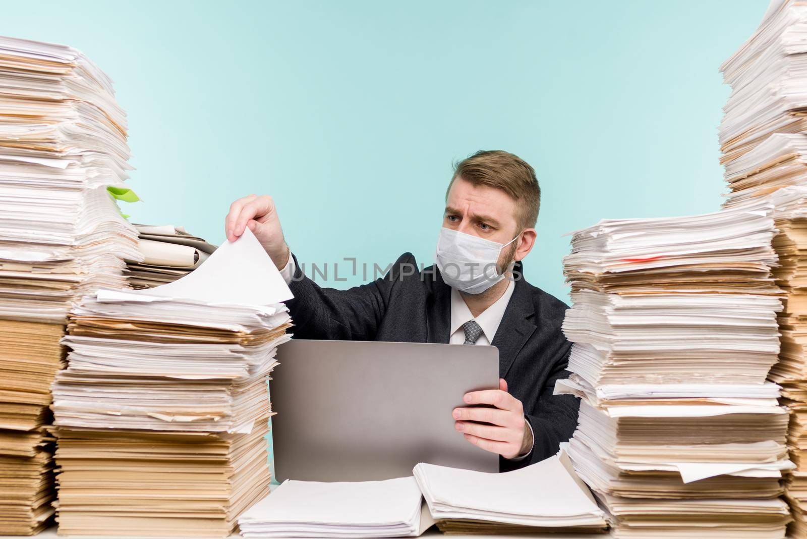 A male accountant or company manager works in an office in a pandemic in view of the accumulated paper work. A protective medical mask is on the face. On the desktop are large stacks of documents. - image