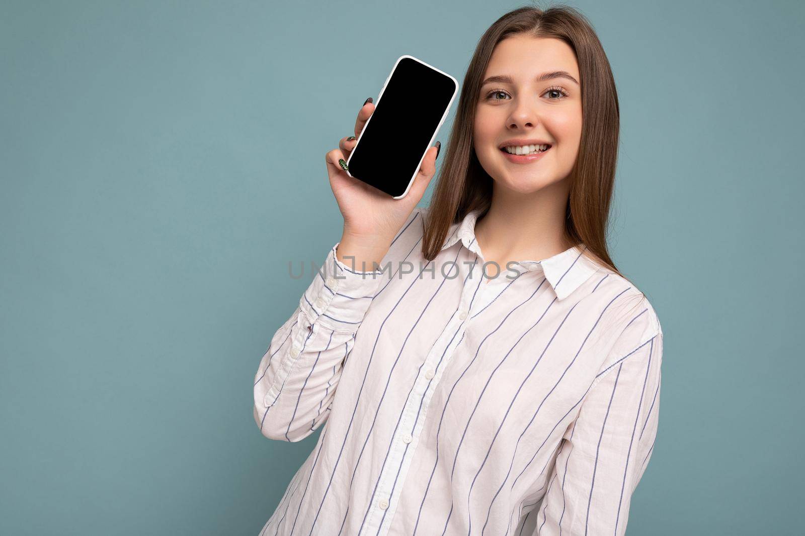 Photo of beautiful smiling young woman good looking wearing casual stylish outfit standing isolated on background with copy space holding smartphone showing phone in hand with empty screen display for mockup looking at camera.