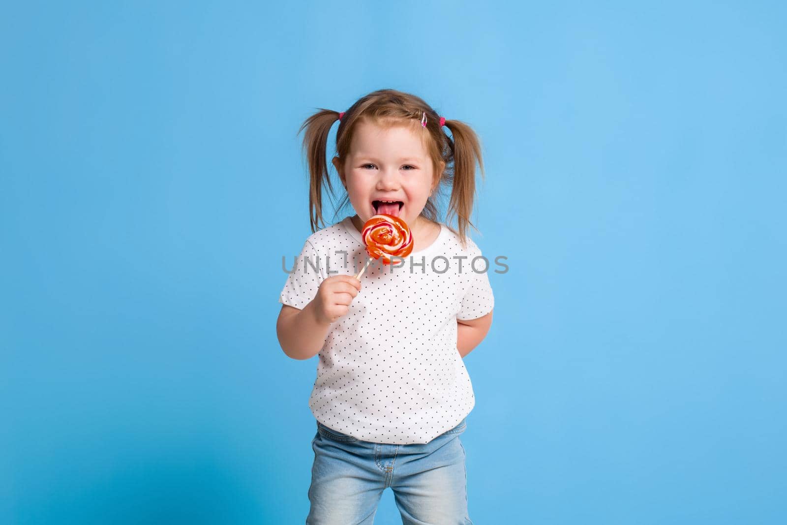 Beautiful little female child holding huge lollipop spiral candy smiling happy isolated on blue background. In children loving sweet and caramel concept