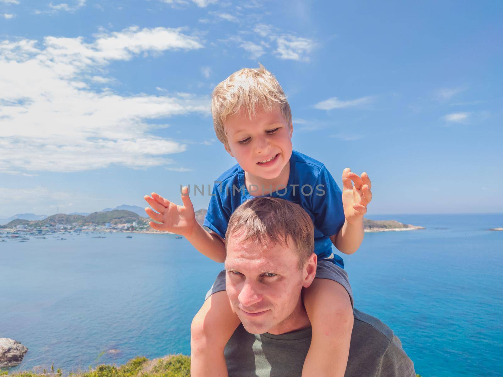 Front boy son laugh sitting on fathers shoulders. No photoshop sun on skin. Sea, clouds, island background. Funny photo, happiness lifestyle, Father's Day, love parenthood, family holiday concept by nandrey85