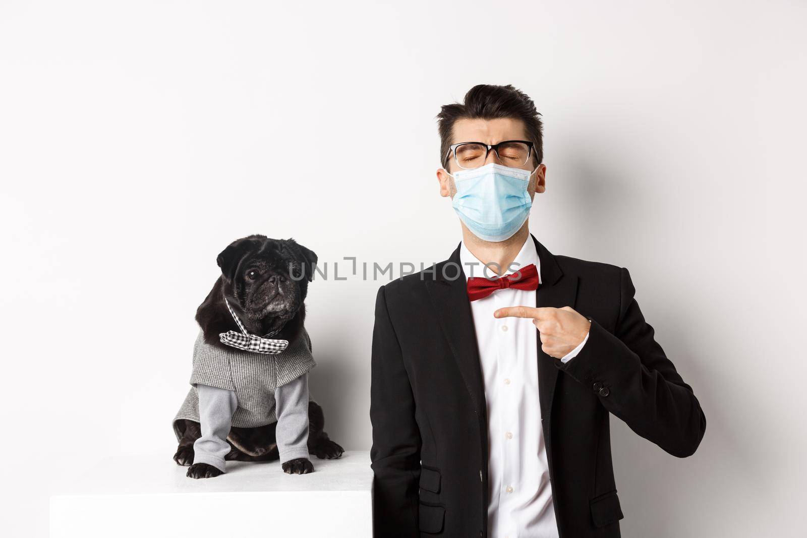 Coronavirus, pets and celebration concept. Disappointed young man in face mask and suit, pointing finger at cute black pug dog wearing party costume, standing over white background.