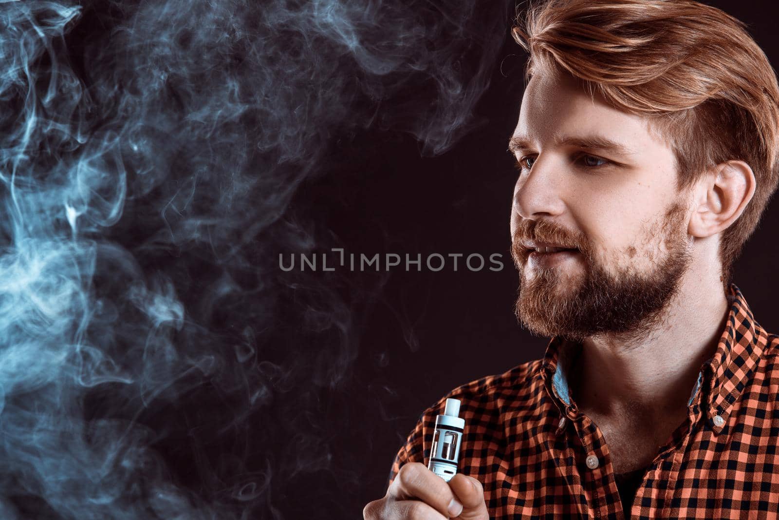 young man wearing a plaid shirt smokes an electronic cigarette on a black background. close-up