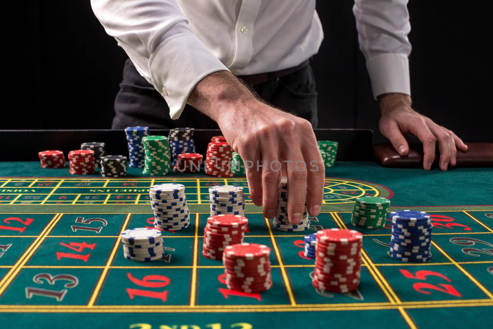 A close-up vibrant image of green casino table with roulette, with the hands of croupier and multicolored chips, on black background. Casino. Gambling. Roulette. Betting