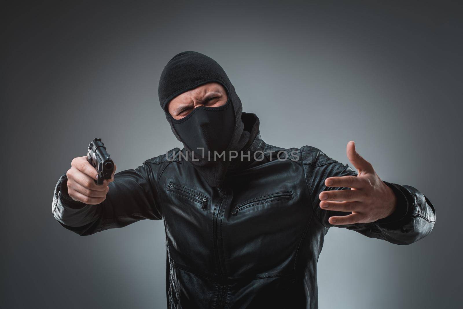 Masked robber with gun, looking into the camera. Studio shot on gray background. Emotions