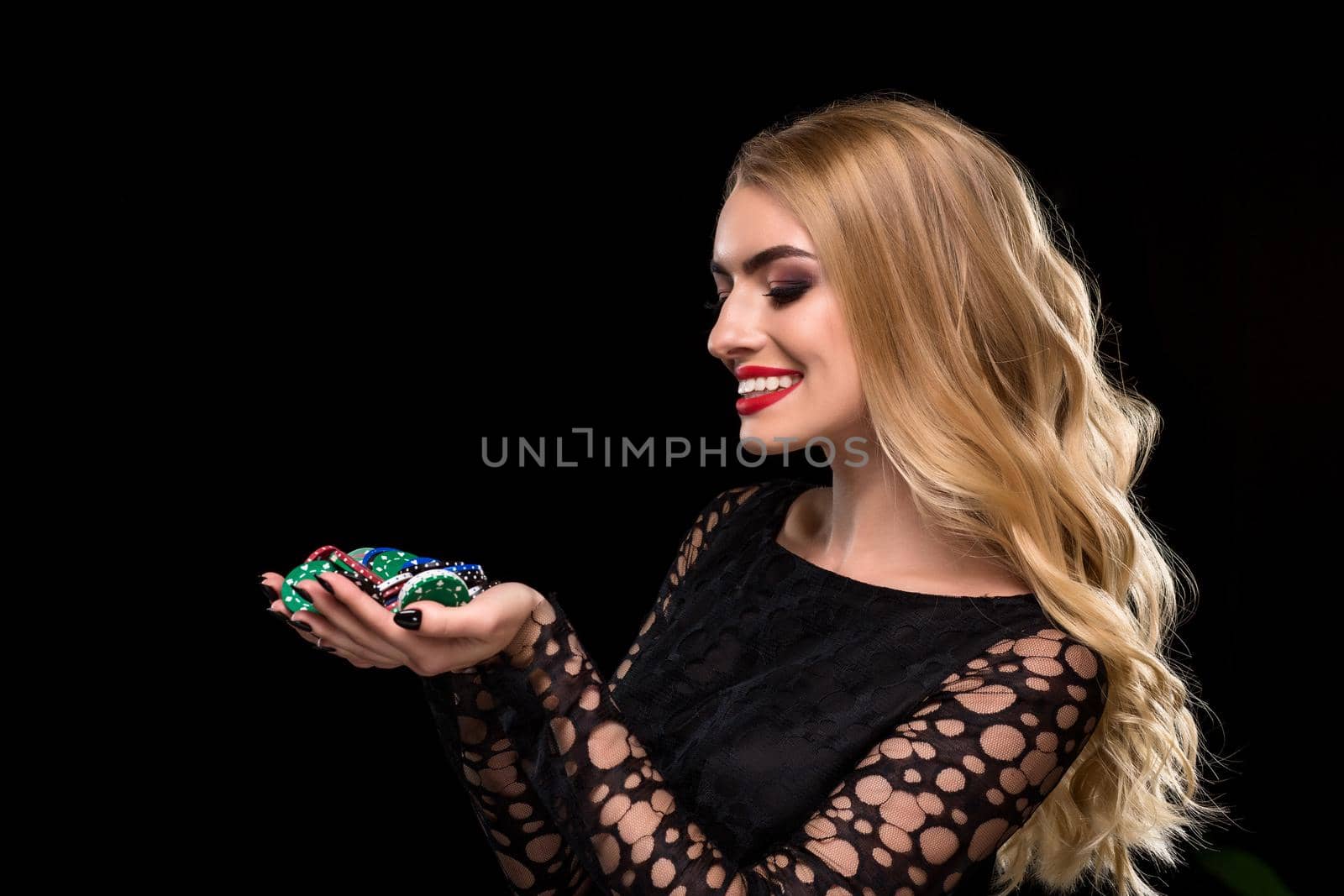 Elegant blonde in a black dress, casino player holding a handful of chips on black background. Poker. Casino. Roulette Blackjack Spin. Caucasian young woman looking at the chips emotions