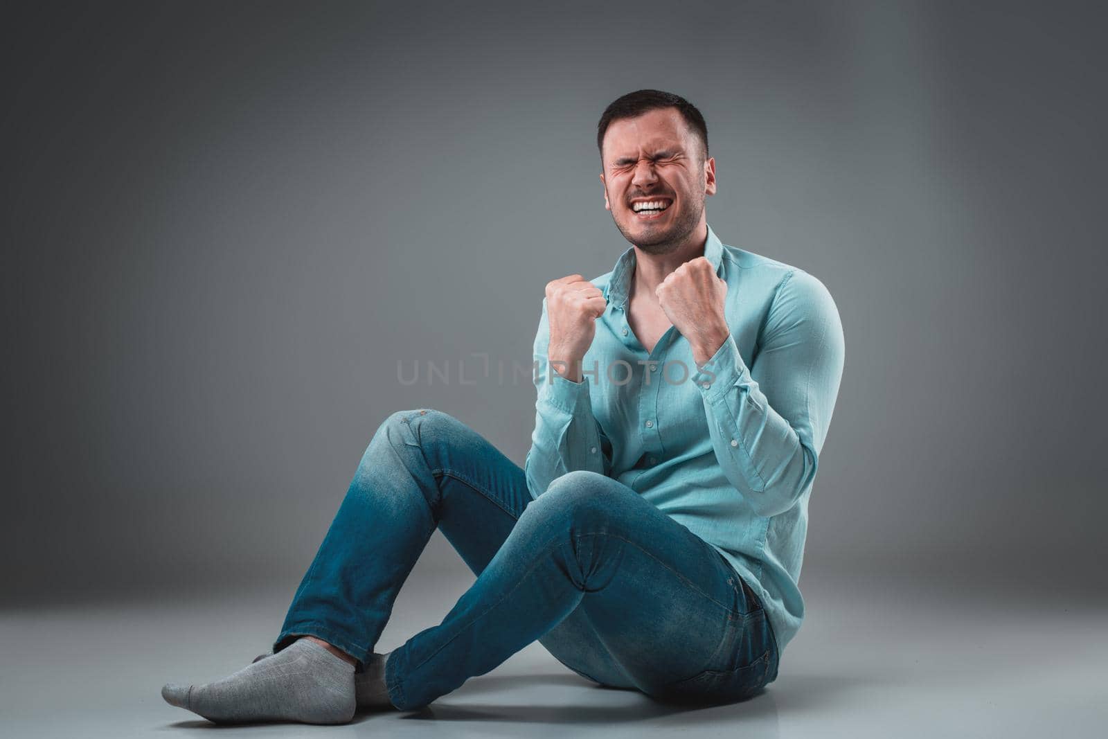 Appealing casual young man sitting on the floor, looking to the camera smiling. Studio shot.