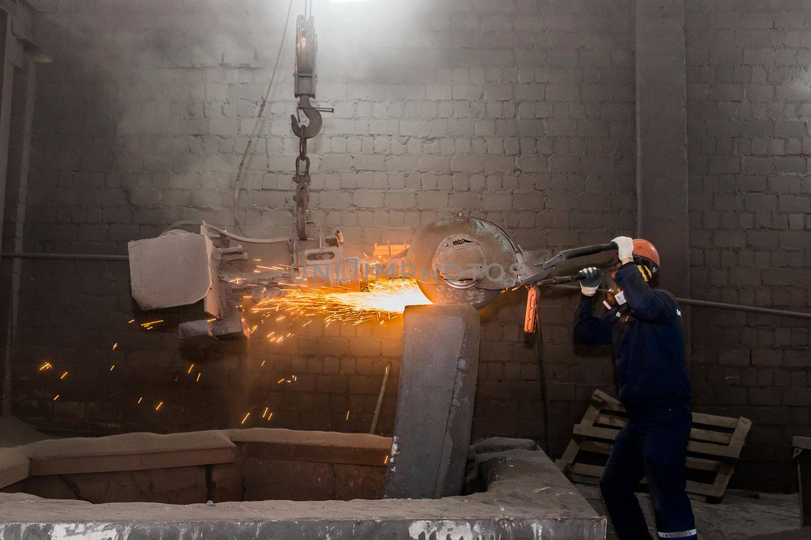 A male worker in a protective helmet, respirator, overalls manages heavy grinding equipment for cast iron concrete tubing with flying sparks in the workshop of an industrial plant by AYDO8