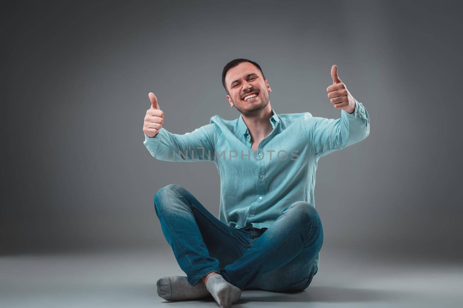 Handsome young man sitting on a floor with raised hands, isolated on gray background. Emotion concept