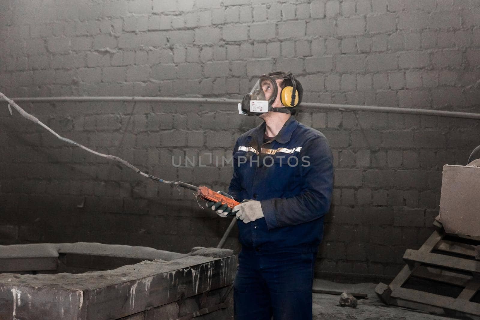 A working man in overalls and a respirator controls the lifting mechanism in the workshop of an industrial plant, with the help of a control panel.