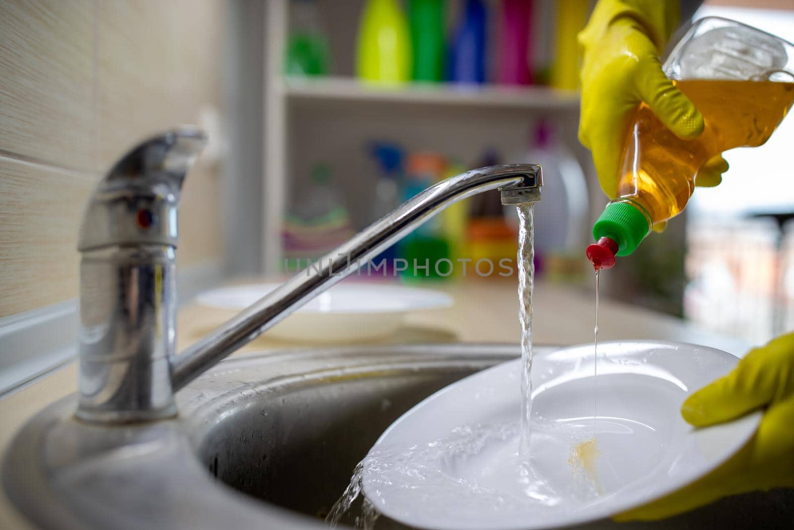 Close up of female hands with gloves pouring liquid detergent on plate for dishwashing under tap water 