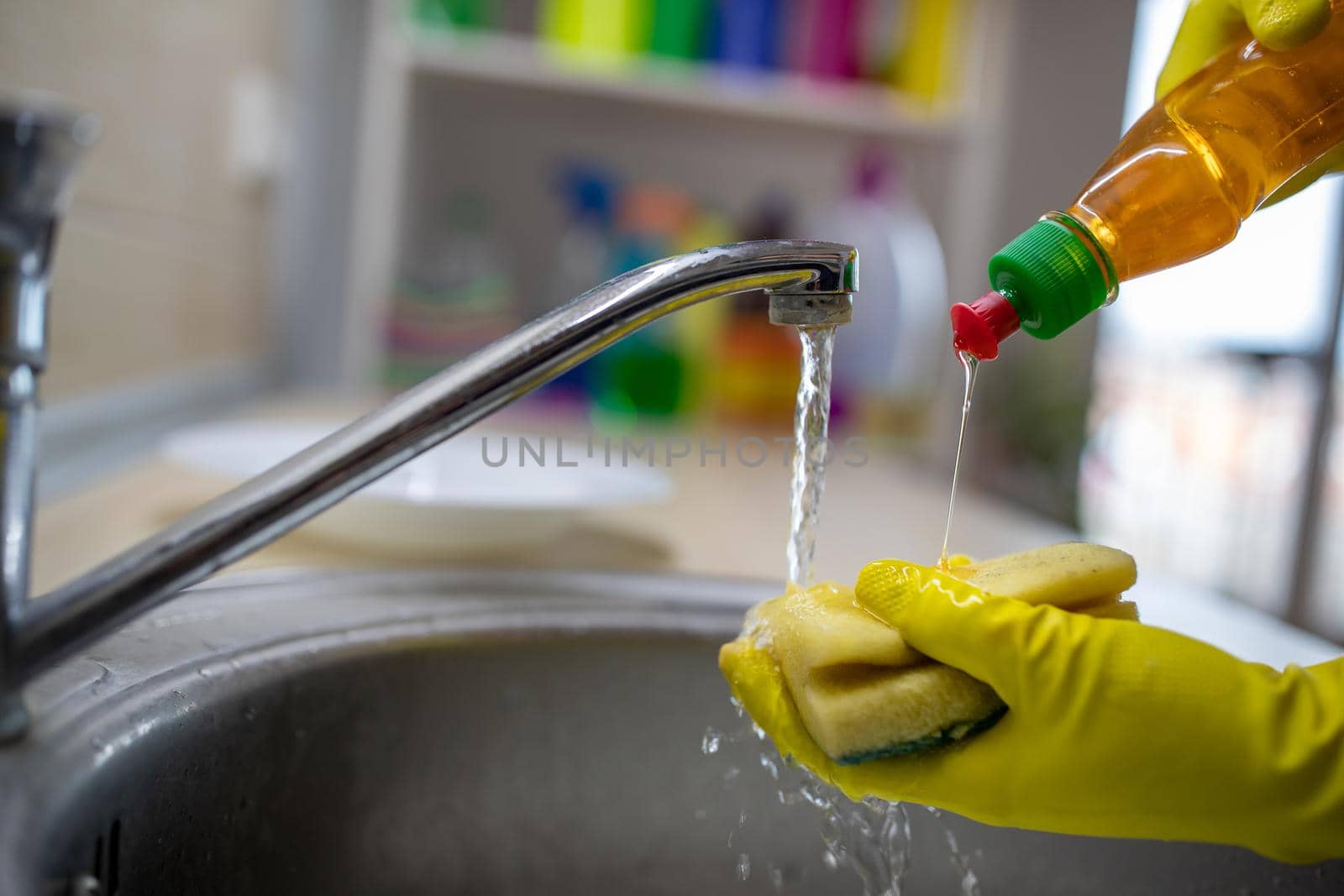 Close up of female hands with gloves holding sponge and pouring liquid detergent above tap water in sing for dish washing