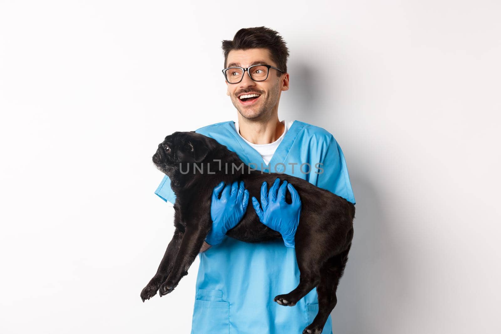 Vet clinic concept. Happy male doctor veterinarian holding cute black pug dog, smiling and looking left, standing over white background.