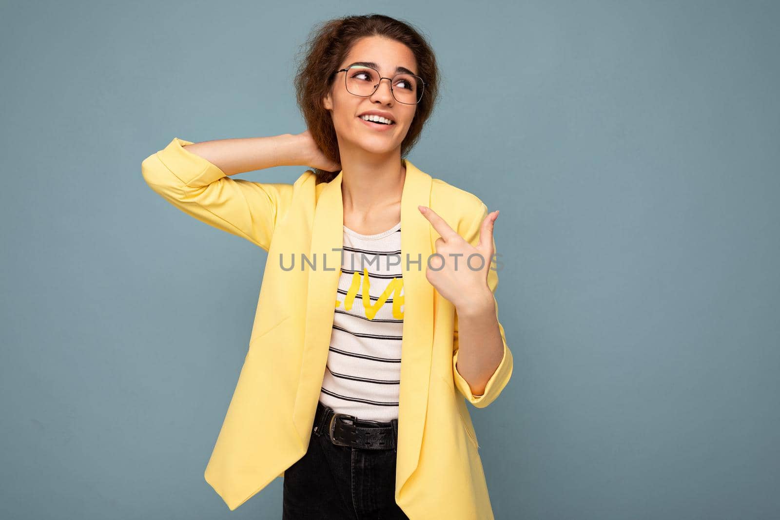 Photo of young positive happy adorable attractive brunette curly woman with sincere emotions wearing stylish yellow jacket and optical glasses isolated on blue background with copy space.