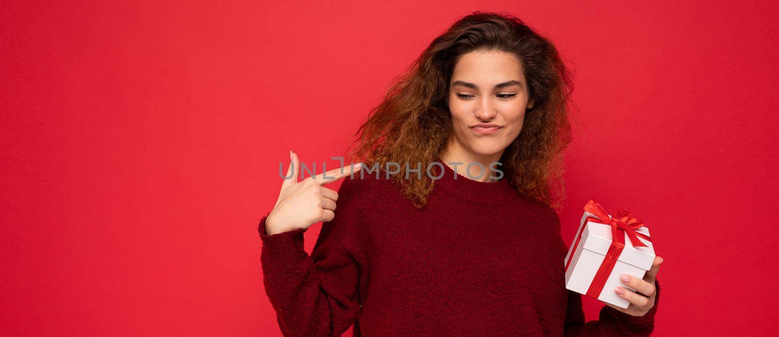 Attractive positive self-confident emotional young brunette curly woman isolated over red background wall wearing red sweater holding gift box looking to the side by TRMK