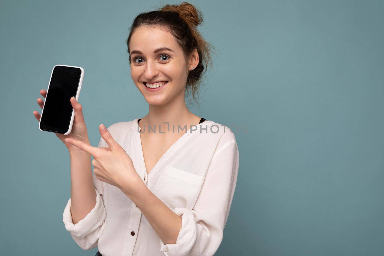 Beautiful positive young blonde curly woman wearing casual white shirt and isolated over blue background wall holding phone and showing smartphone with empty dcreen for mockup looking at camera and pointing finger at device.