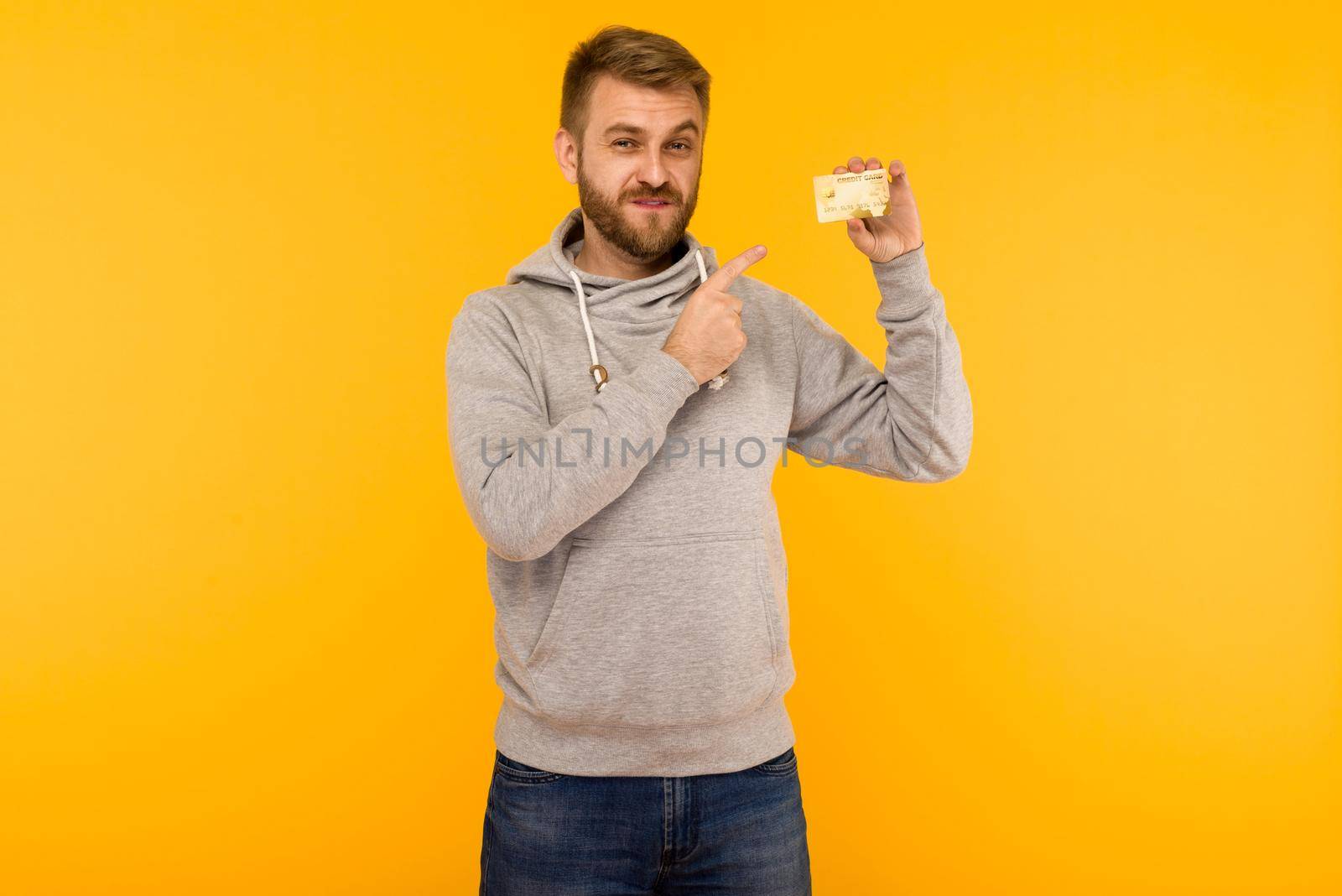 Attractive man in a gray hoodie points a finger at the credit card that is holding in his hand on a yellow background by zartarn