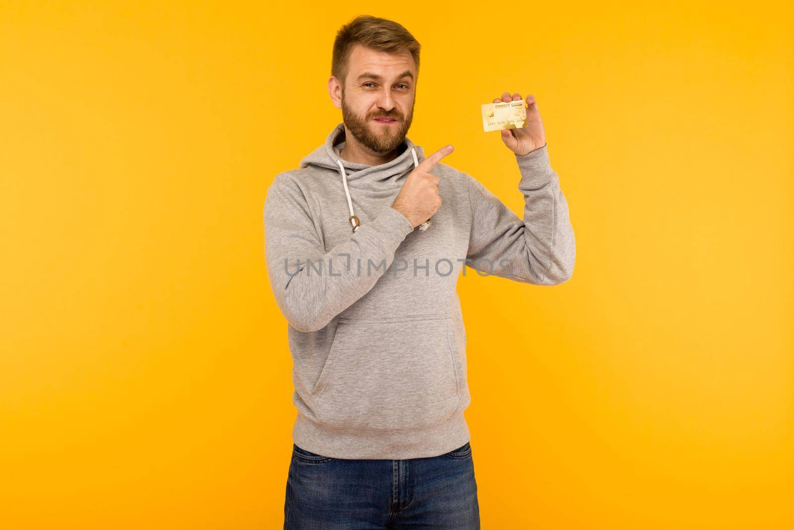 Attractive man in a gray hoodie points a finger at the credit card that is holding in his hand on a yellow background - image