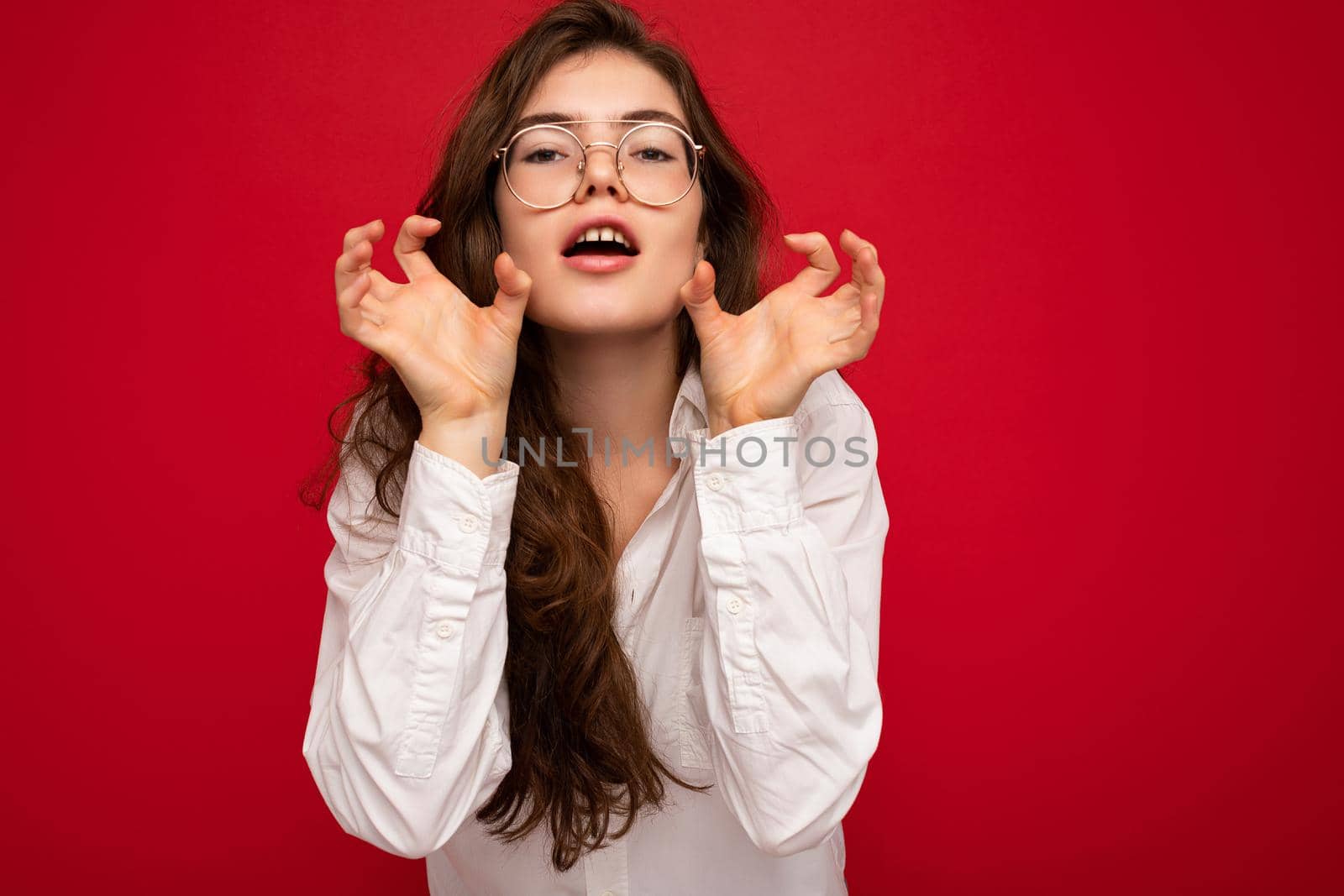 Portrait of young emotional positive sexy beautiful brunette woman with sincere emotions wearing casual white shirt and optical glasses isolated on red background with copy space and making cat claws and growling like animal.