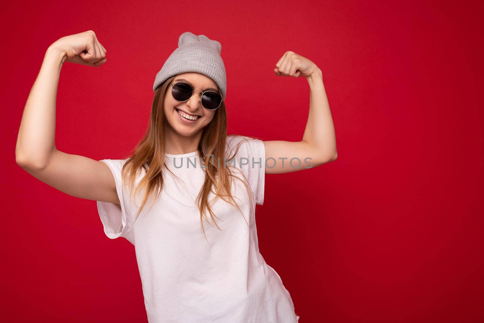 Photo of young positive happy smiling beautiful woman with sincere emotions wearing stylish clothes isolated over background with copy space and showing strong muscles.