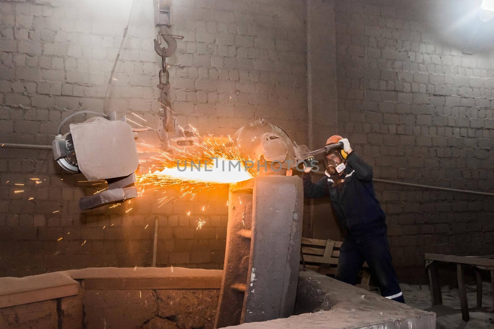 A male worker in a protective helmet, respirator, overalls manages heavy grinding equipment for cast iron concrete tubing with flying sparks in the workshop of an industrial plant.