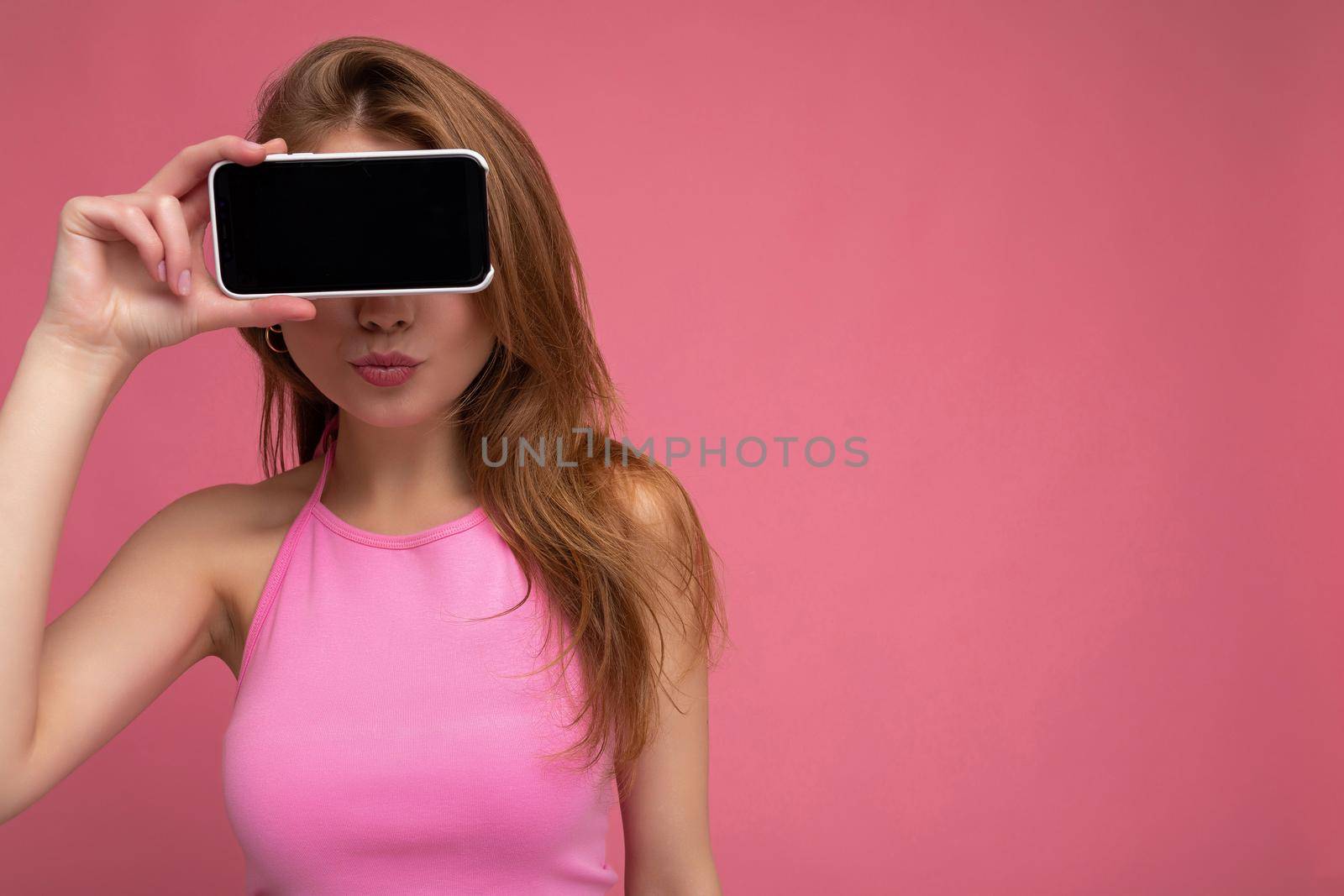 Photo of beautiful positive young blonde woman wearing pink top poising isolated on pink background with empty space holding in hand and showing mobile phone with empty display for mockup.