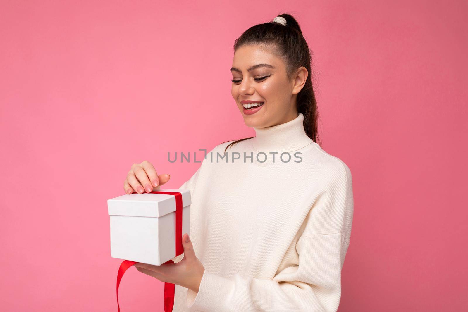 Charming positive surprised young brunette woman isolated over pink background wall wearing white sweater holding gift box and unboxing present looking at box.