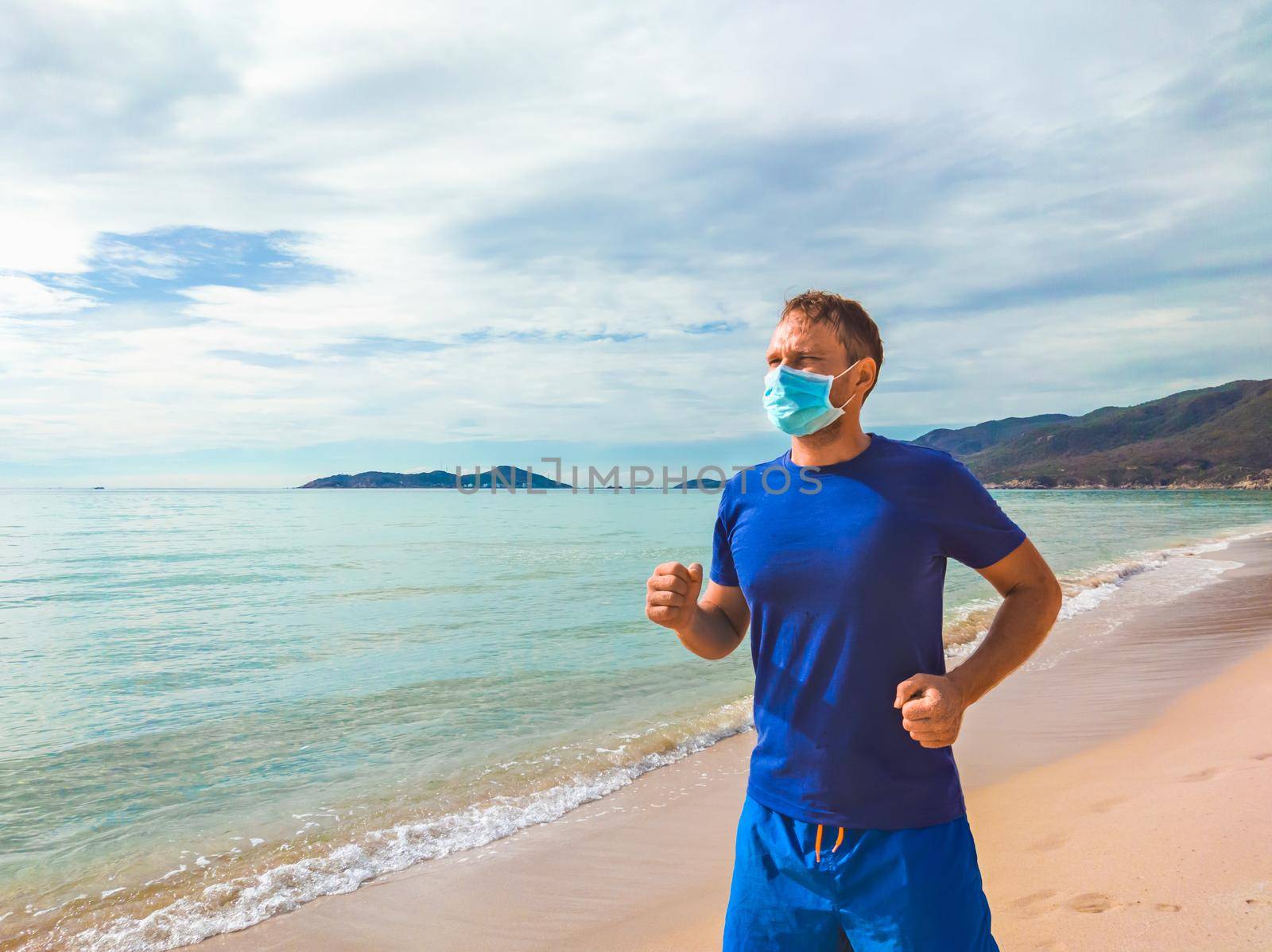 Coronavirus COVID 19. Young beautiful man doing sport performing workouts exercises and running near sea in the morning during quarantine. Active life in surgical sterilizing face mask protection by nandrey85