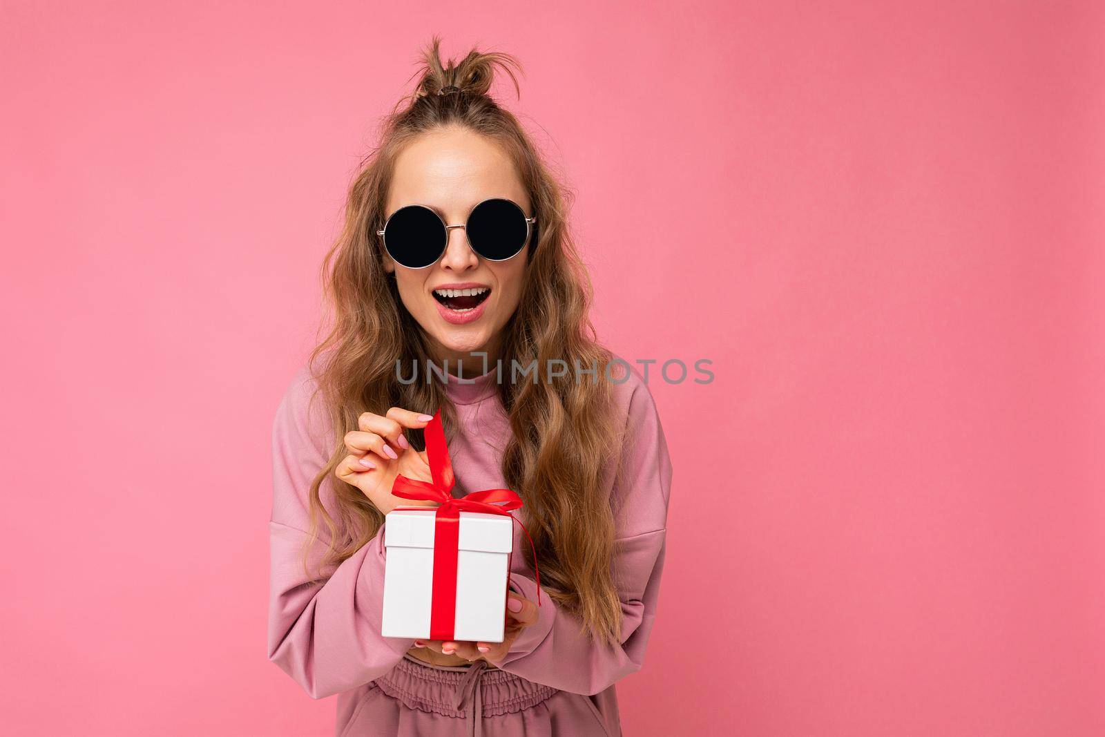 Pretty surprised happy young blonde curly female person isolated on pink background wall wearing stylish pink sport clothes and sunglasses holding gift box looking at camera. Free space