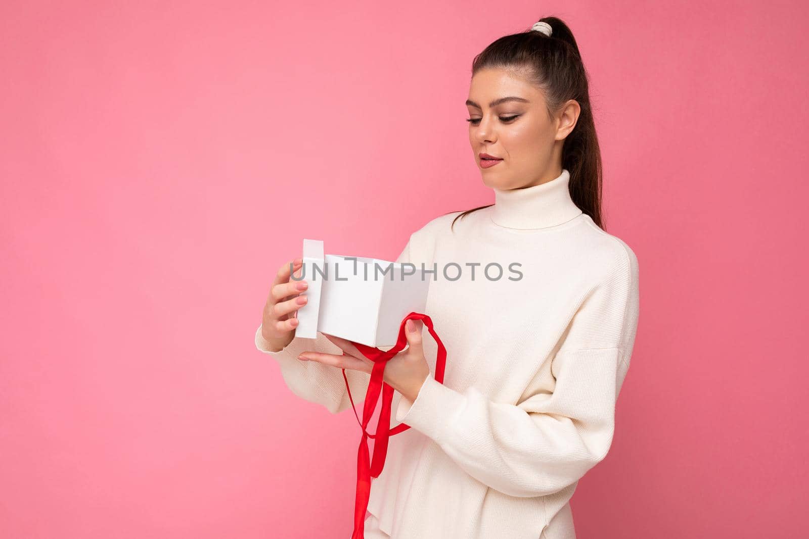 Attractive happy amazed young brunette woman isolated over pink background wall wearing white sweater holding gift box and unboxing present looking at camera.